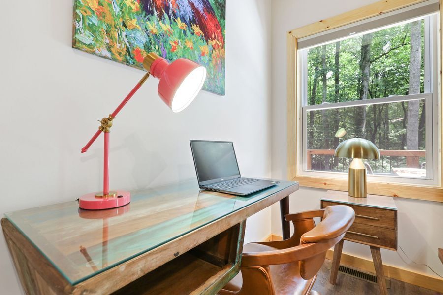 Desk with laptop, pink lamp, and chair beside a window overlooking a wooded area.