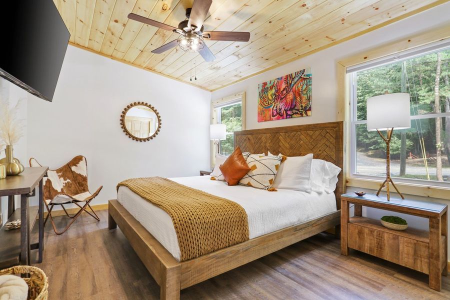 Bedroom with wooden bed frame, ceiling, and nightstand. Features artwork and cowhide chair.