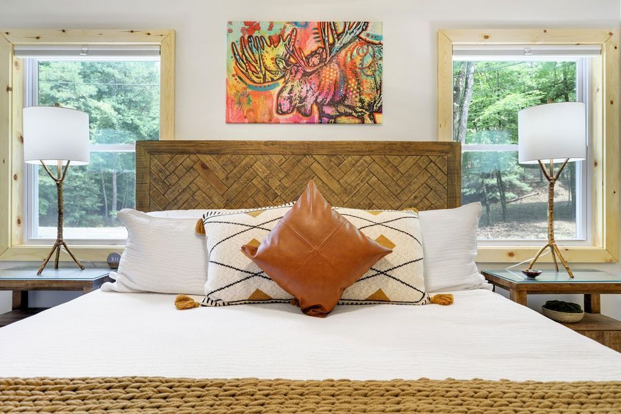 Bedroom with bed, wooden headboard, two windows, artwork above, lamps, and nightstands.