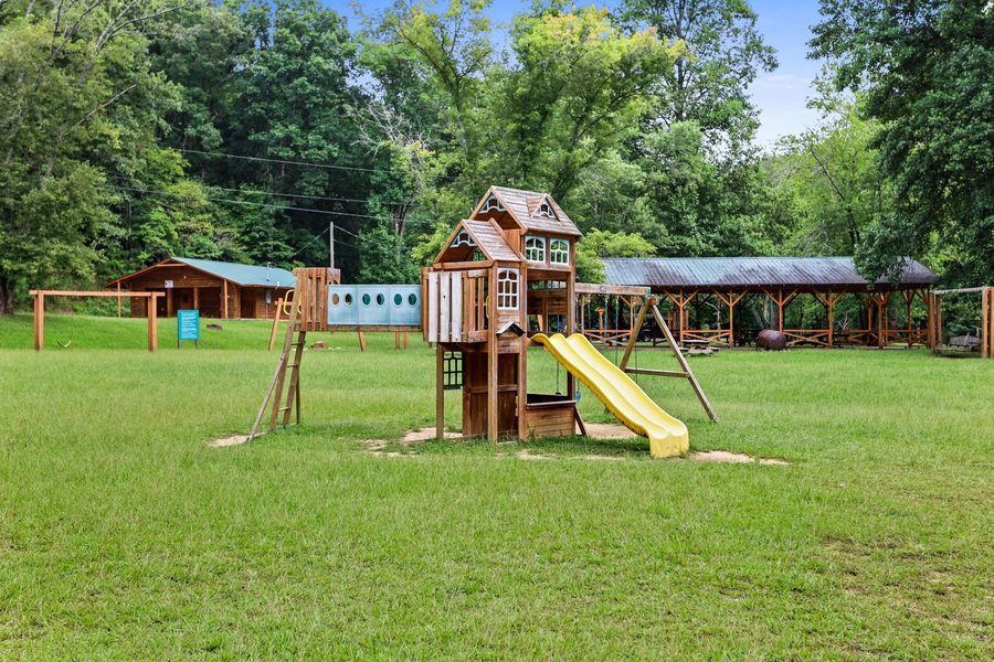 A wooden playground with a slide and swingset on a grassy field, surrounded by trees and a covered structure.