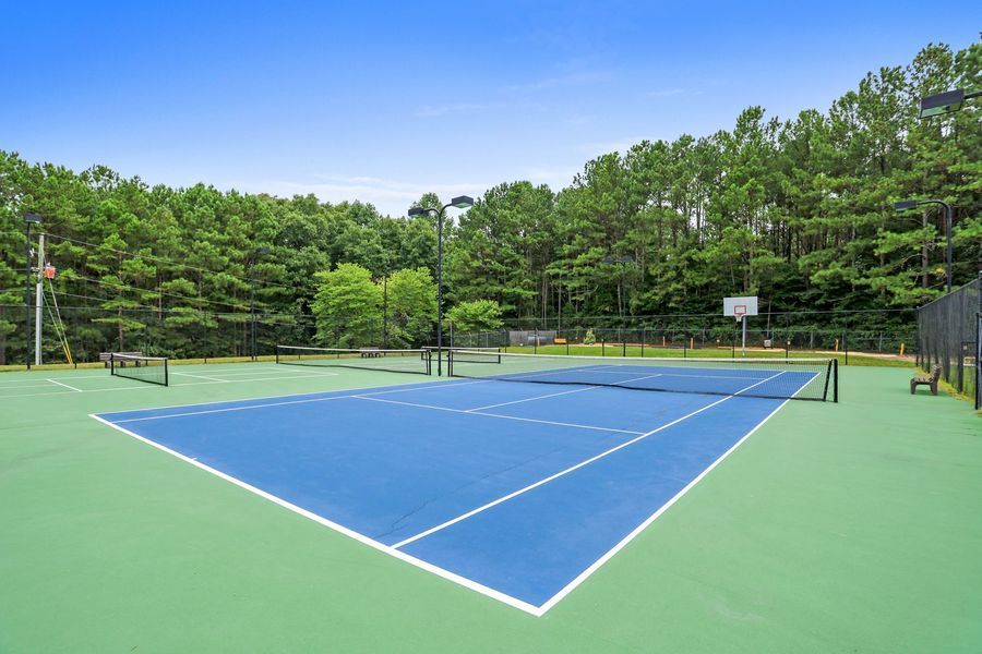 Blue tennis court with net and green surroundings under blue sky.