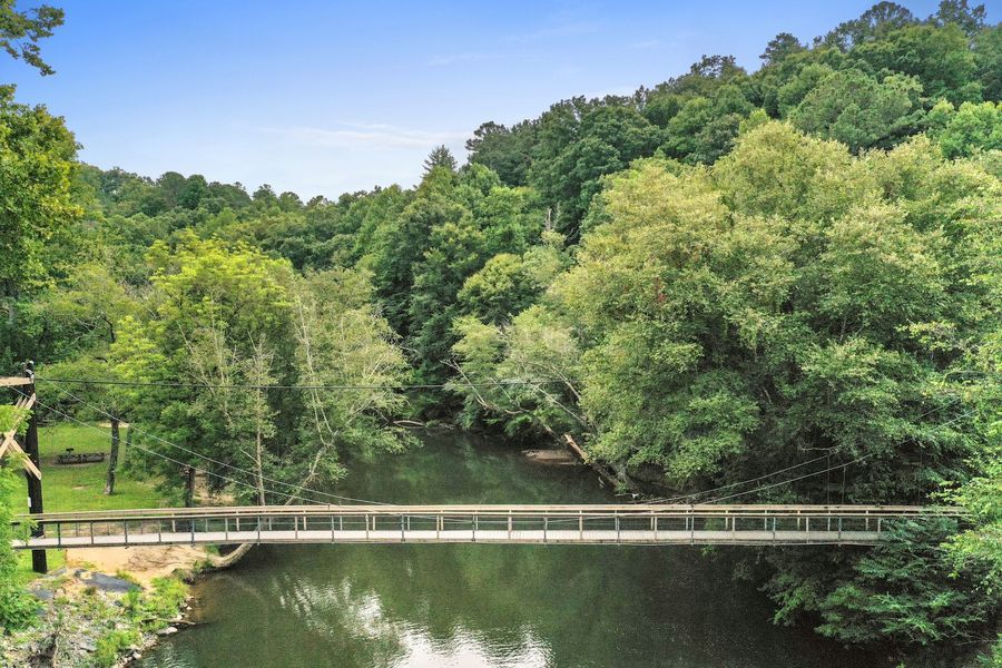 A wooden bridge spans a calm, tree-lined river. Lush green trees surround the water under a blue sky.