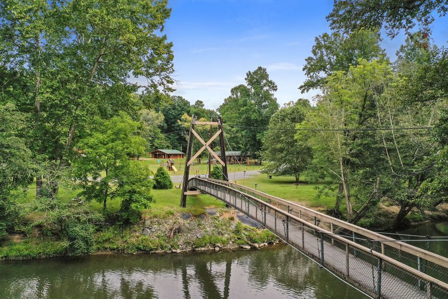 A bridge spans a river, leading to a green grassy area surrounded by trees.