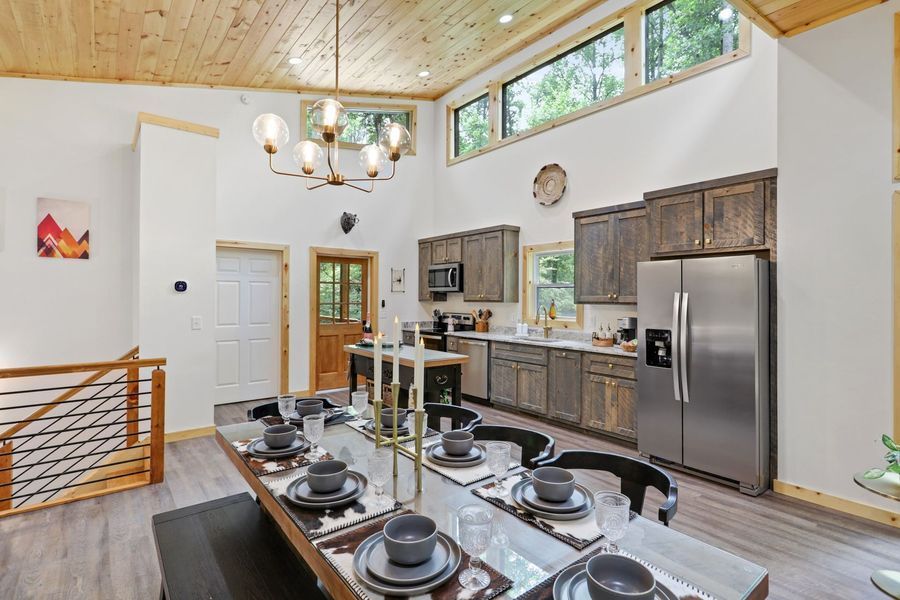 Spacious cabin interior with kitchen, dining table, and tall windows. Wood ceiling, grey cabinets, and modern chandelier.