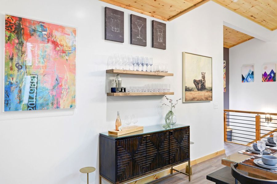 A dining area with a colorful painting, a credenza with glassware, and a wooden ceiling.