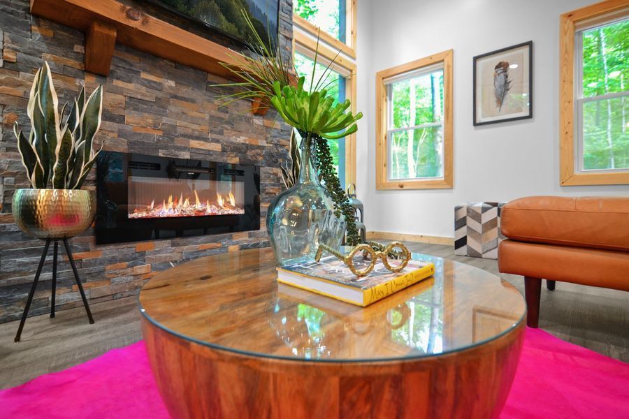Living room with fireplace, round wooden coffee table, pink rug, tan leather armchair, windows with a view.