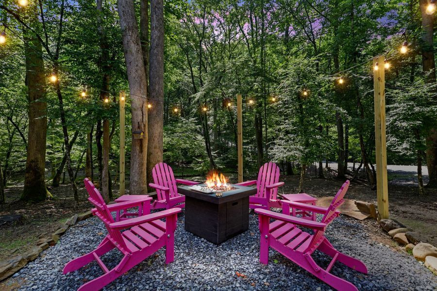 Pink Adirondack chairs around a fire pit in a wooded area, illuminated by string lights.