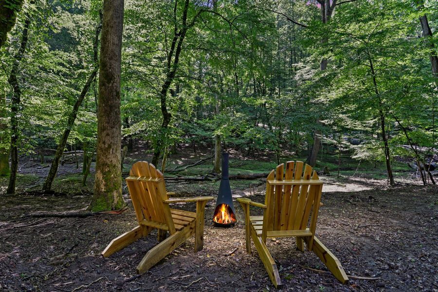 Two wooden Adirondack chairs face a fire pit in a forest setting.