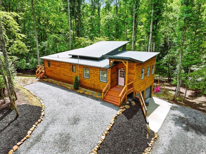 Cabin nestled in a forest with a gravel driveway and dark mulch border. Wooden structure with red stairwell.