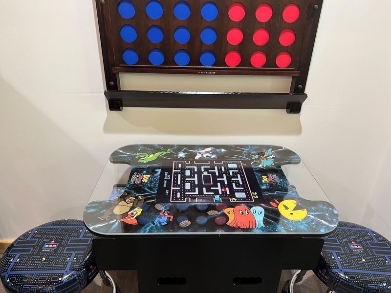 Arcade game table with Pac-Man theme and Connect 4 board above it, with stools.