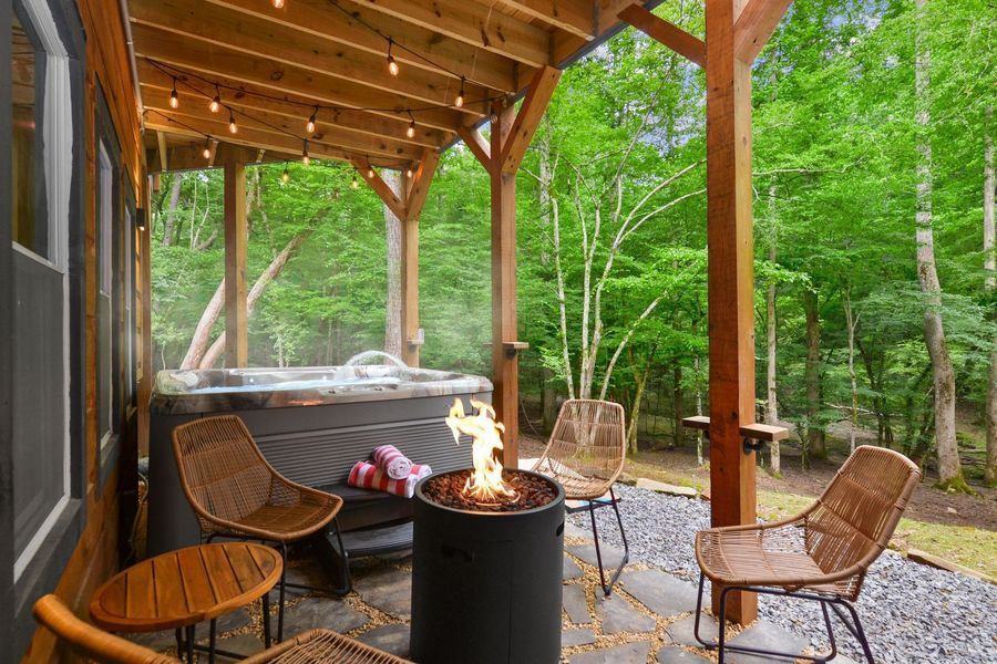 Covered patio with hot tub, fire pit, and seating in a forest setting.