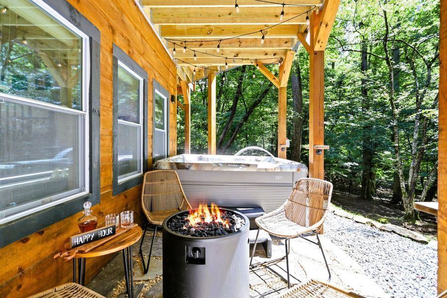 Cozy cabin patio with hot tub, fire pit, chairs, and string lights in a wooded setting.