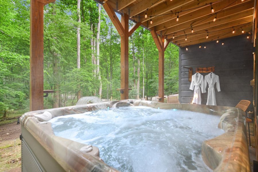 Hot tub on covered porch in wooded area; two white robes hanging.