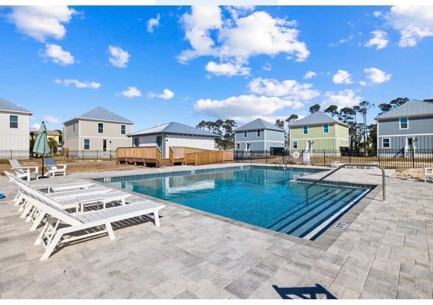 Pool surrounded by lounge chairs and houses under a bright blue sky with fluffy clouds.