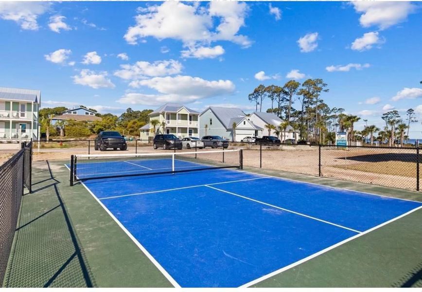 Blue pickleball court with net, surrounded by fence and buildings, under blue sky.