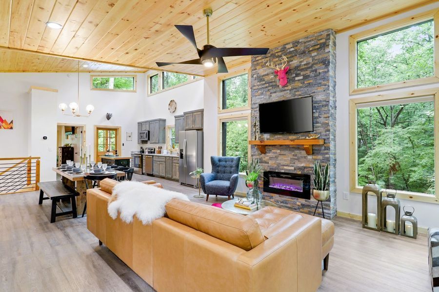 Spacious cabin interior with a modern kitchen, fireplace, and expansive windows with outdoor views.