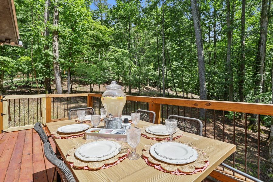 Outdoor dining table set with plates, glasses, and a lemonade dispenser on a deck surrounded by trees.
