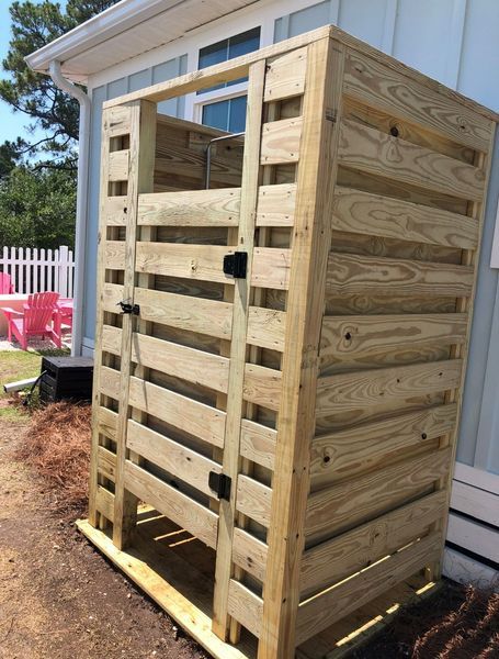 Wooden outdoor shower stall next to a light blue house with a white picket fence in the background.