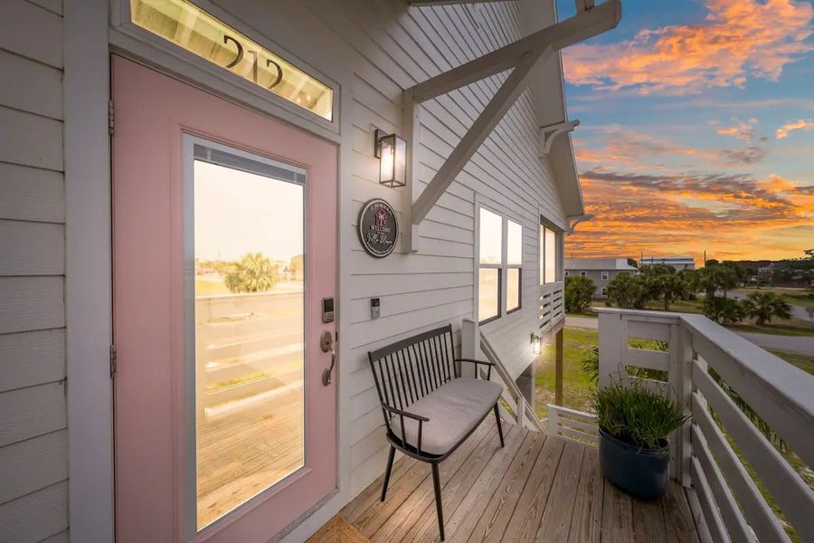 Exterior view of a house with a pink door, balcony with a bench, and sunset.