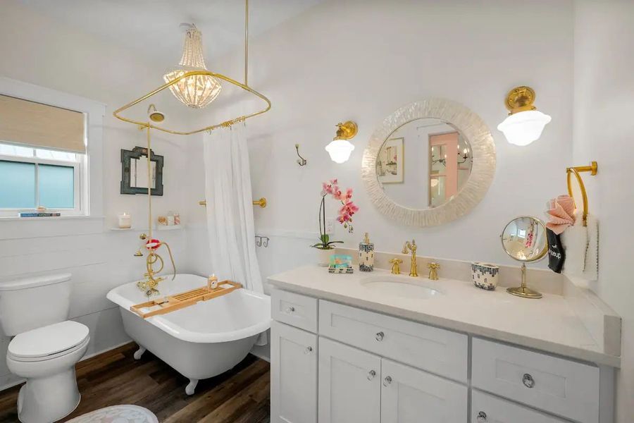 Bright bathroom with clawfoot tub, white vanity, gold fixtures, and a decorative chandelier over the shower.