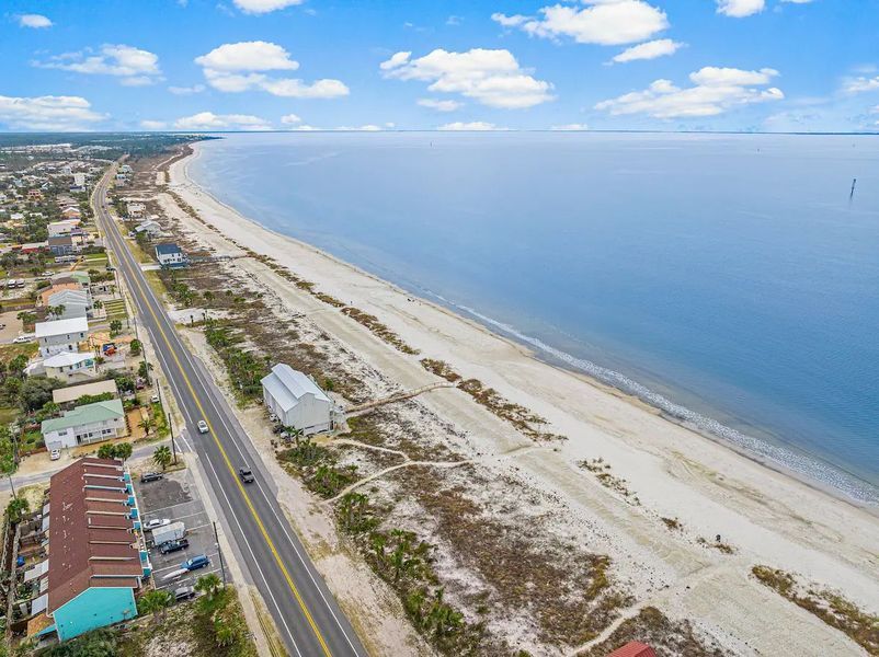 An aerial view of a coastal road, beach, and calm blue water under a partly cloudy sky.