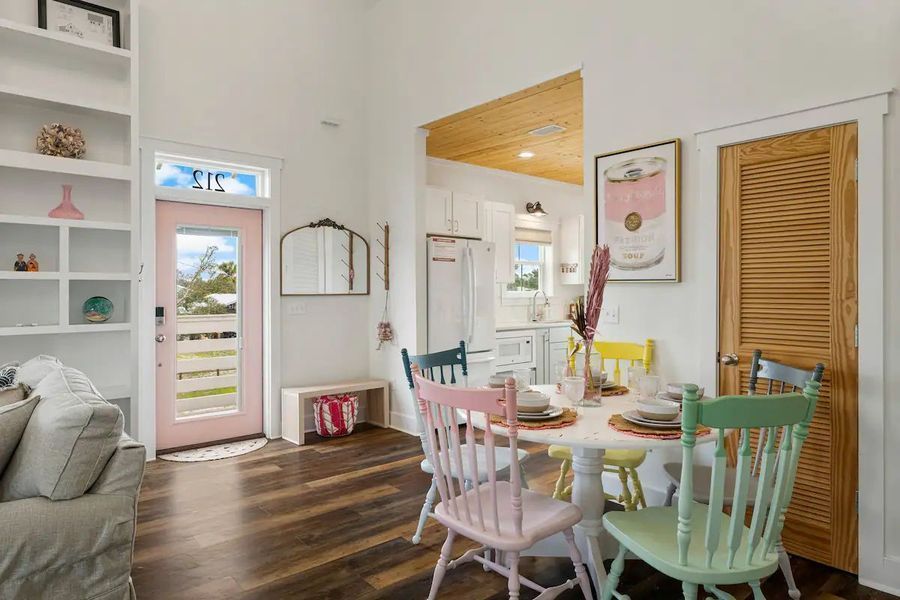 A light-filled dining room with colorful chairs. Pink door, wooden floors, and open to a white kitchen.