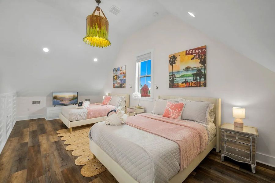 Bedroom with two beds, art, and window. Light wood floors, white walls. Beds have pink bedding and decor.