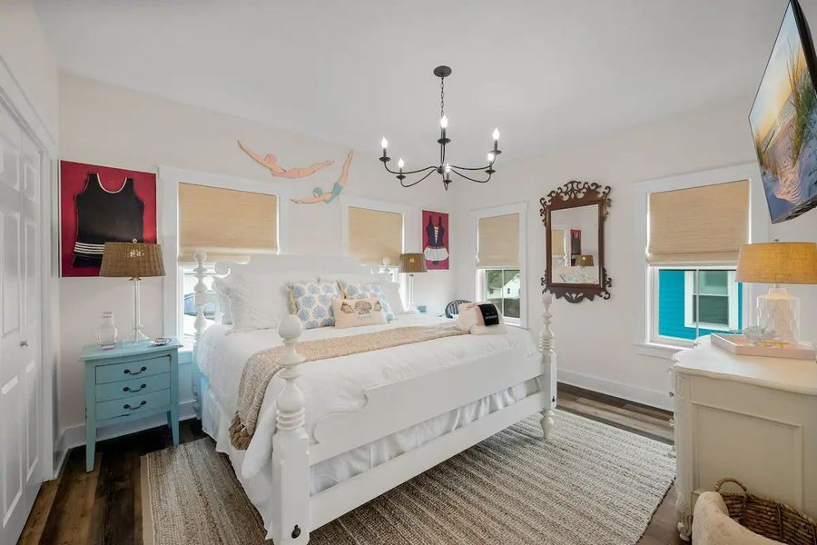 Bright bedroom with white bed, blue nightstand, chandelier, art, and windows.
