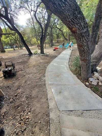 A concrete path curves through a park with trees. Blue chairs and a red object are at the path's end.