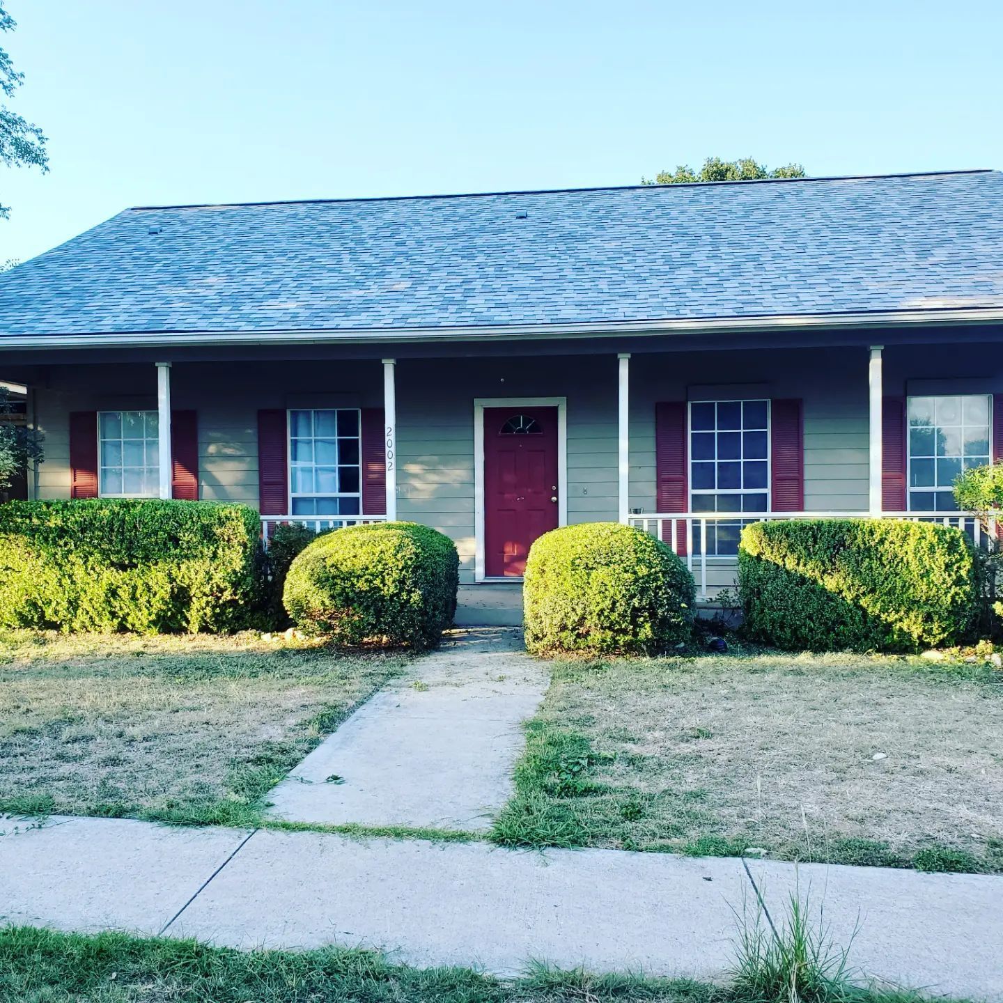 Tan ranch-style house with red door and shutters, green bushes, and concrete walkway on a sunny day.