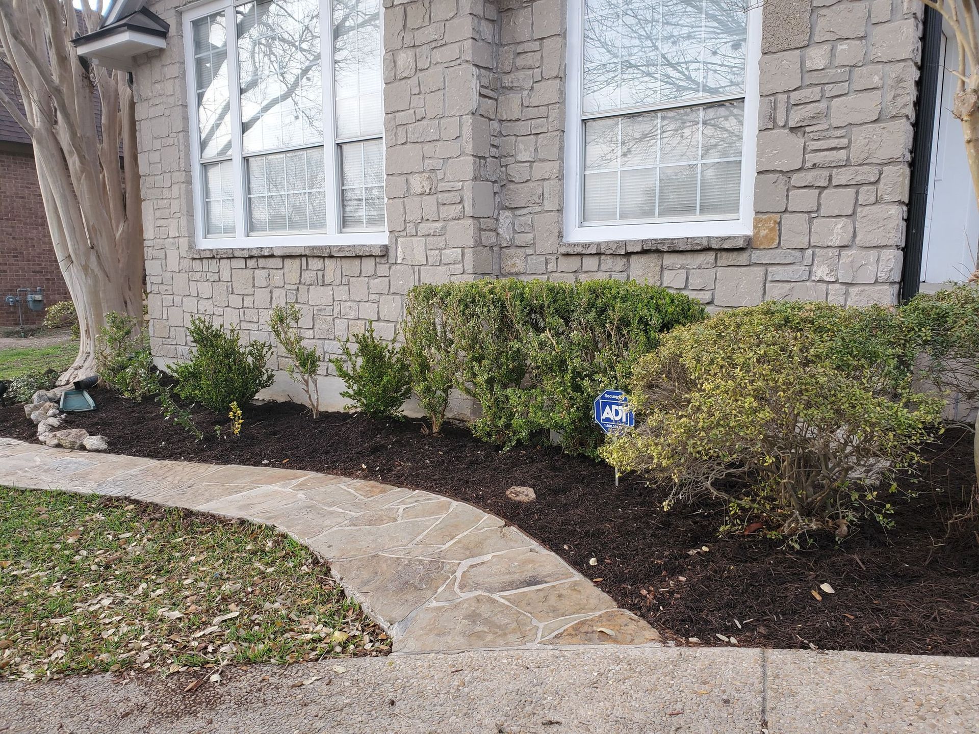 Stone-faced house with white-framed windows, walkway, bushes, dark mulch.