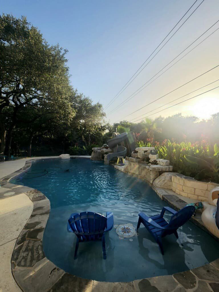Swimming pool with waterfall feature, two blue chairs, and a table. Sunlit, outdoor setting.