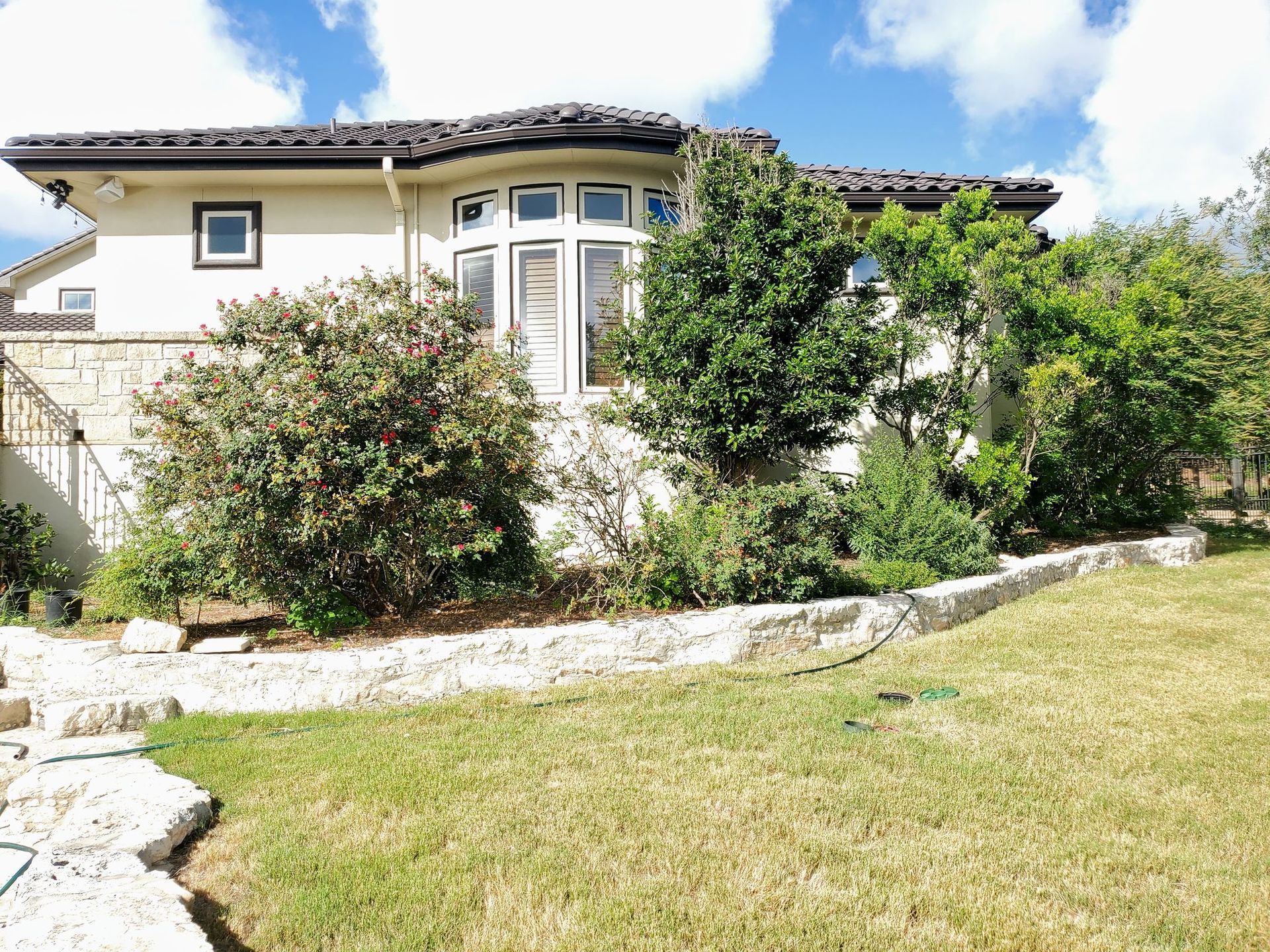 House with curved front windows, surrounded by green shrubs and a grassy lawn under a blue sky.