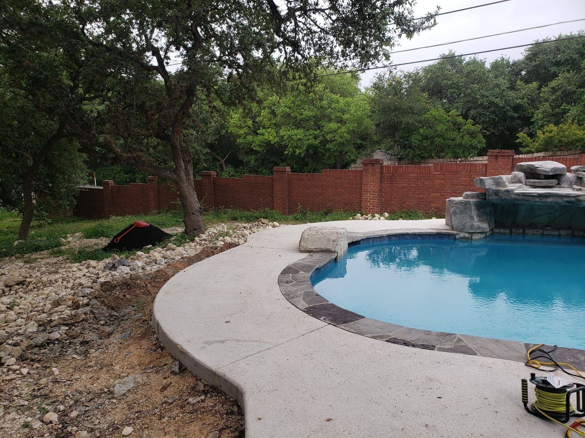 Poolside view with concrete walkway, brick wall, blue water, and rocky landscaping.