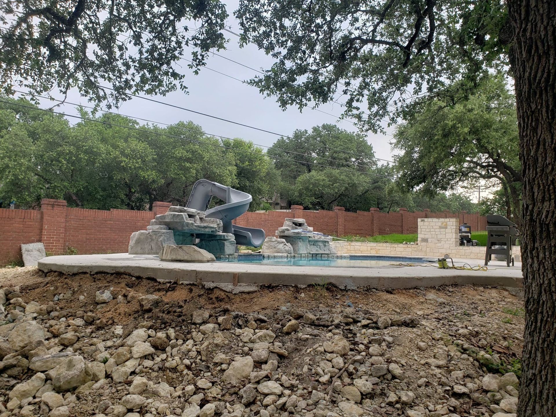 A water feature with blue rock design sits on a concrete pad, surrounded by dirt and a red brick wall, under a cloudy sky.