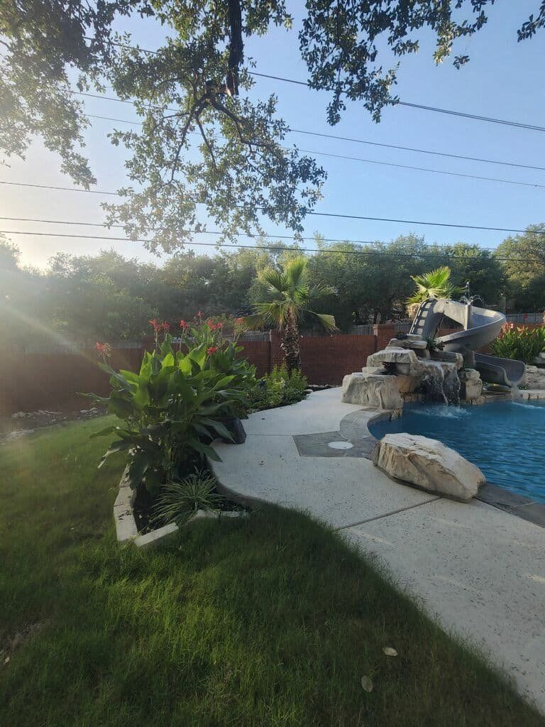 Backyard pool with rock waterfall and slide. Green lawn, trees, and blue sky.