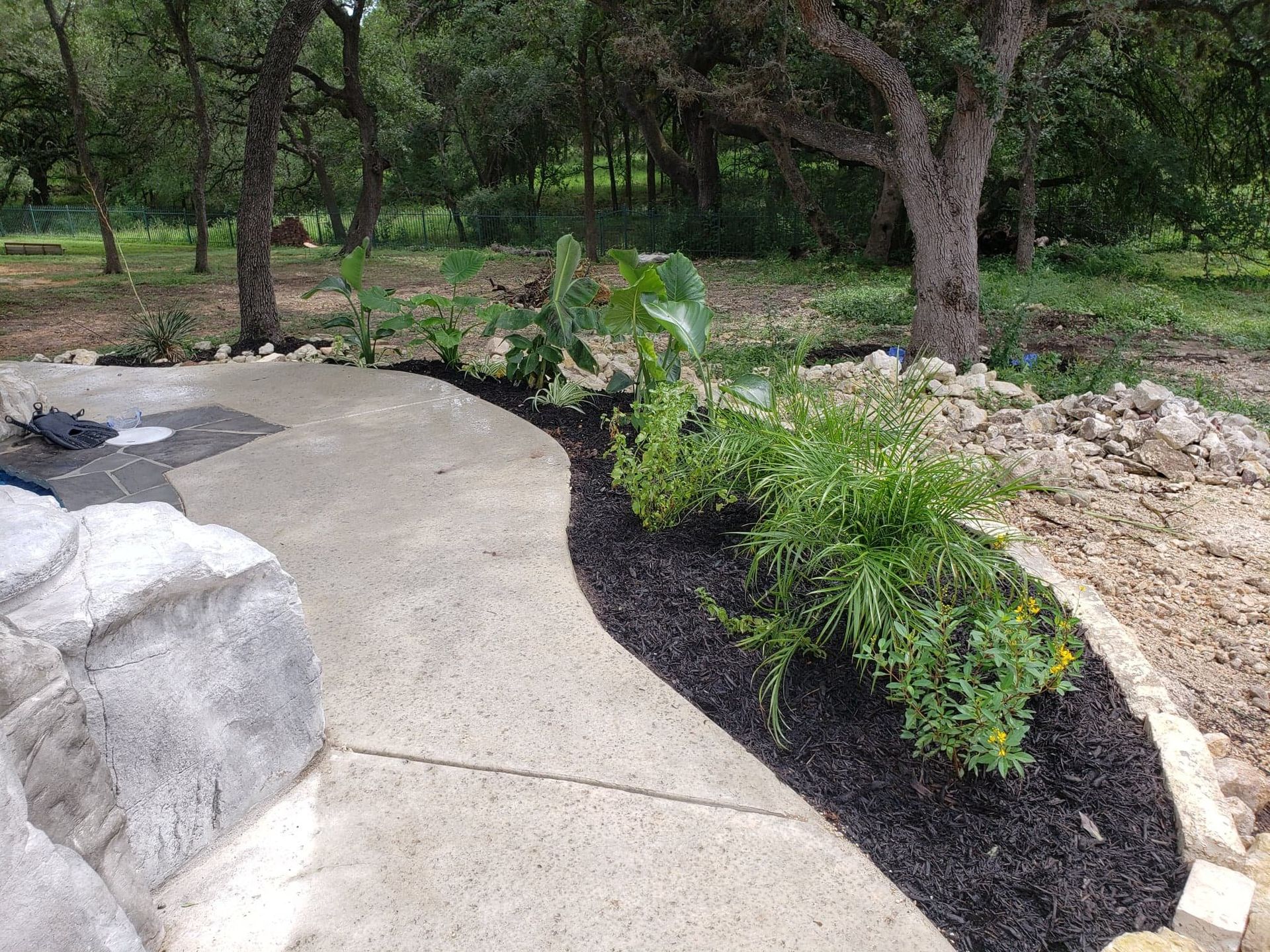 Concrete walkway curves past a garden bed with plants, mulch, and a textured wall. Trees and foliage are in the background.