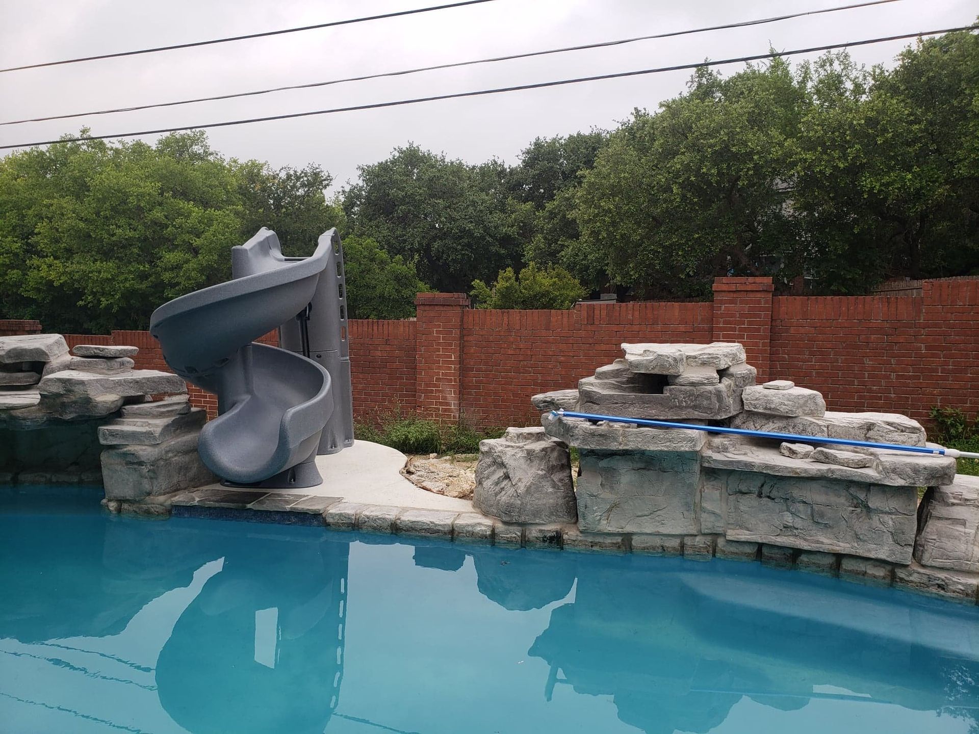 Pool with a gray slide and rocky features. Brick wall and trees in the background.