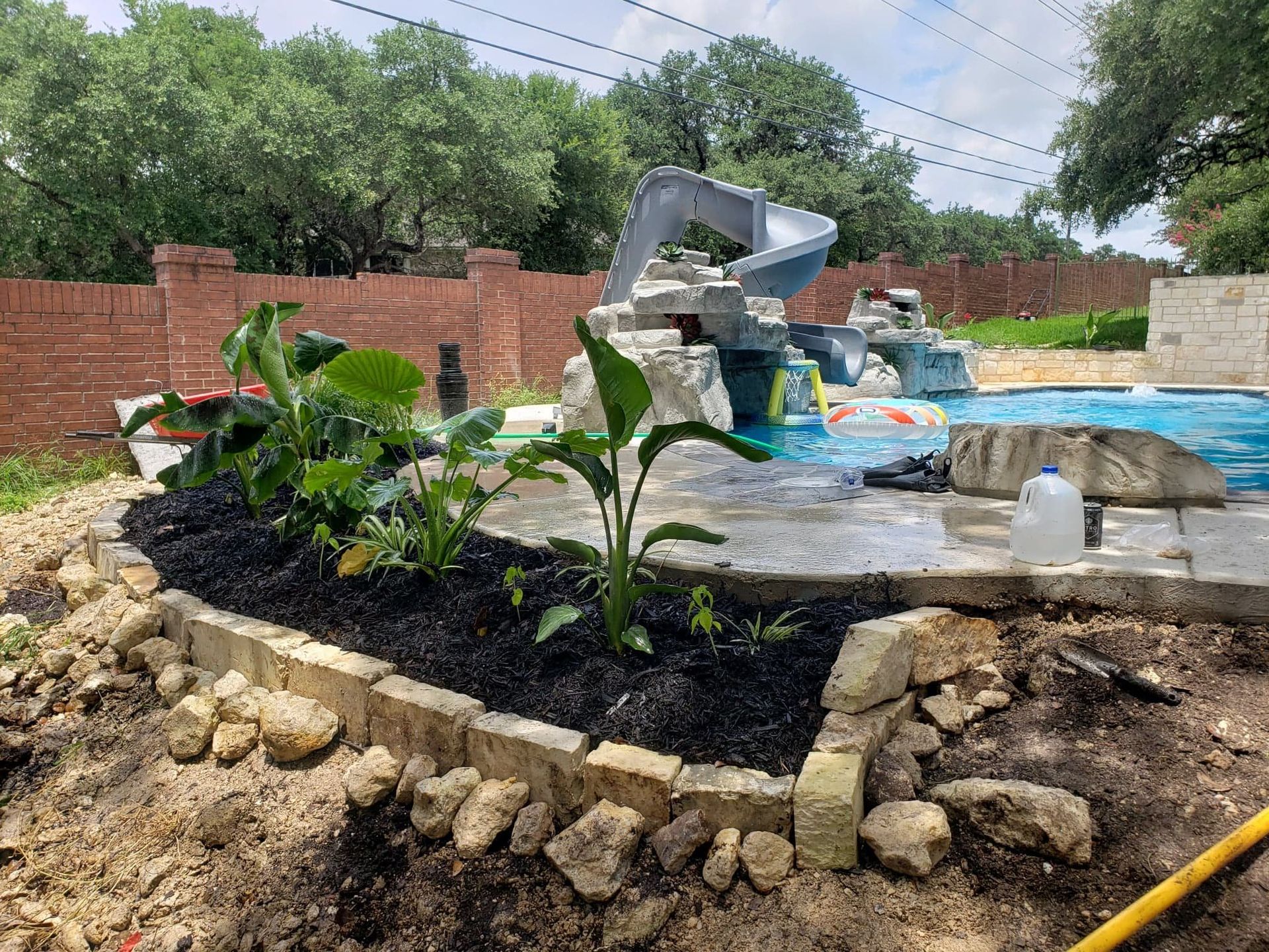 Pool area with a water slide, plants, and brick wall. Freshly mulched planting bed.