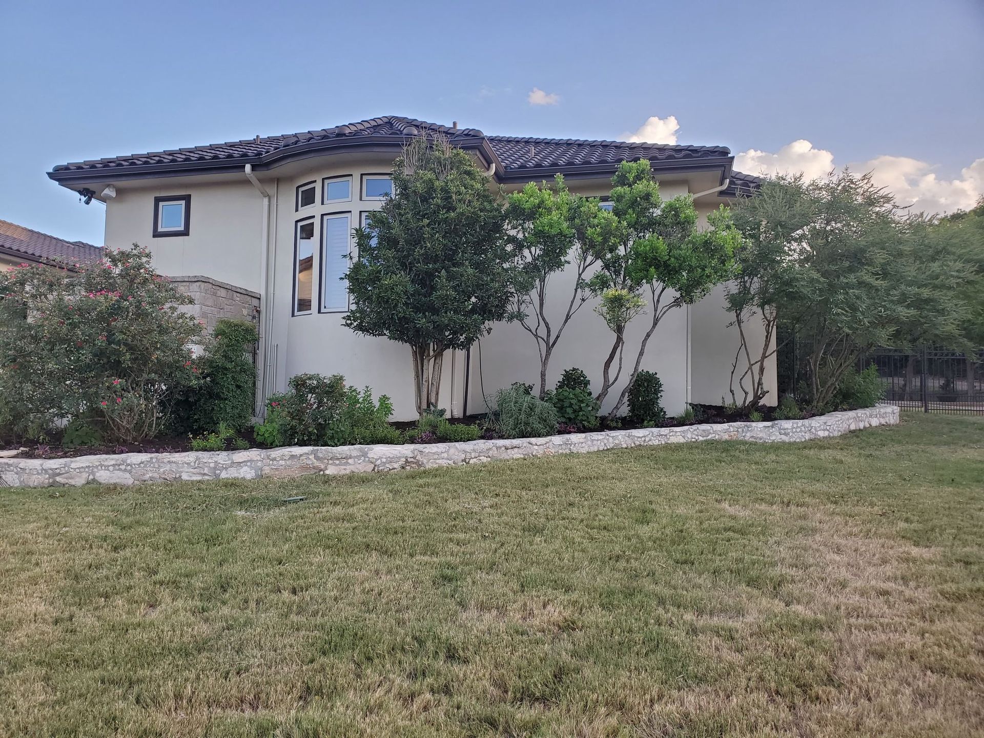 Beige stucco house with a tile roof, surrounded by landscaping and a grassy lawn.
