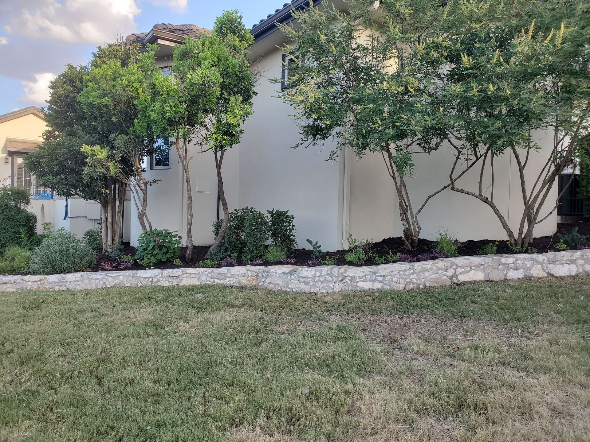 A house with a light-colored stucco exterior, trees, and a stone retaining wall in front.