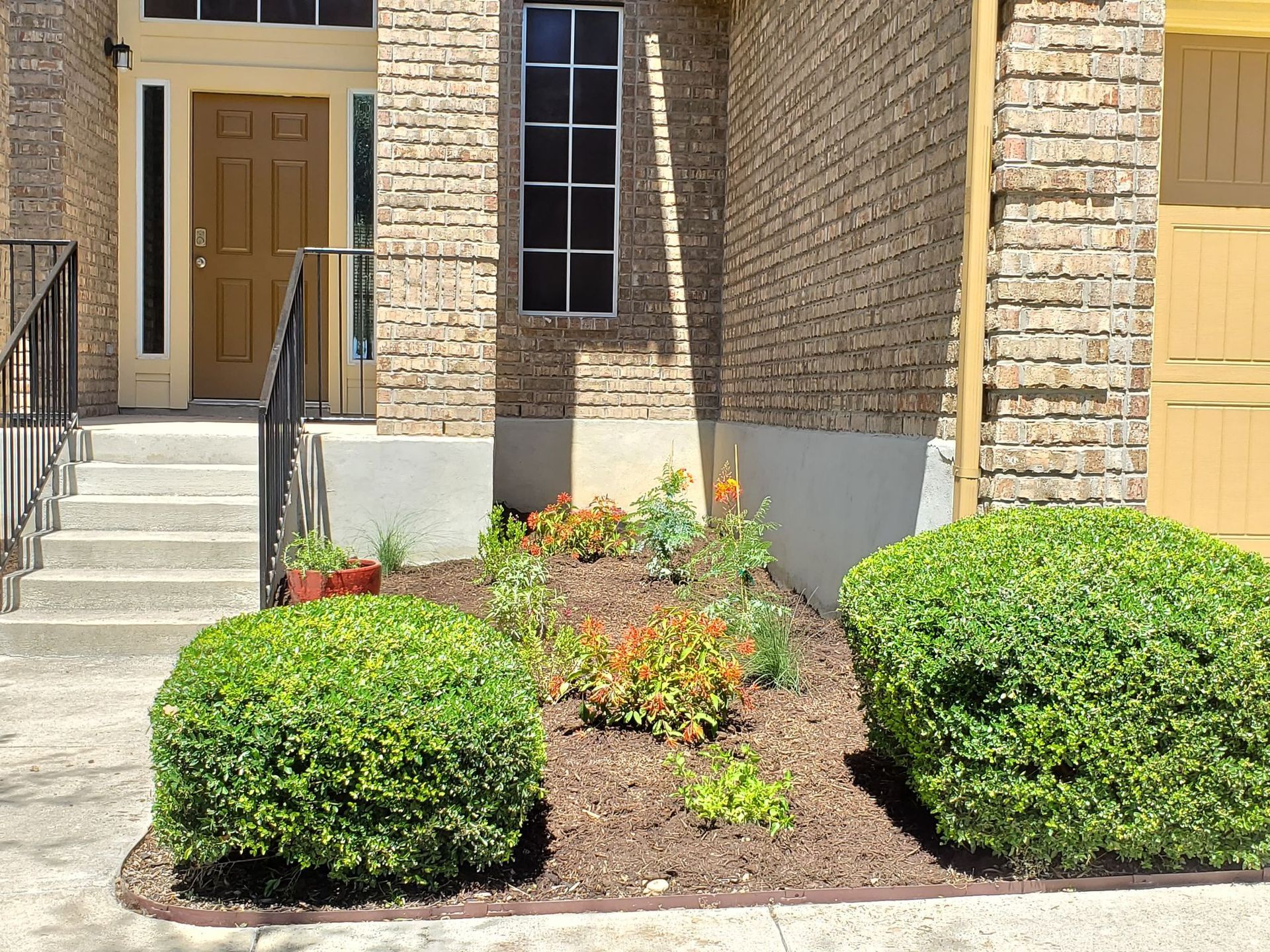 A house exterior with a front door, steps, and a flower bed with bushes and plants.