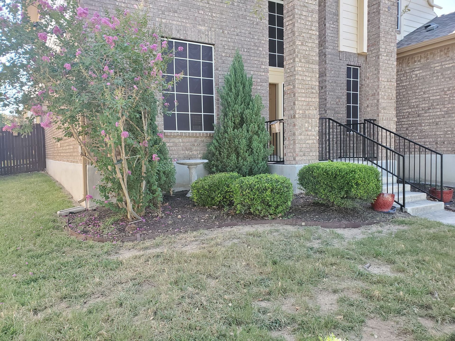 Front of a brick house with landscaping; lawn in front, trees, bushes, and a black metal railing.