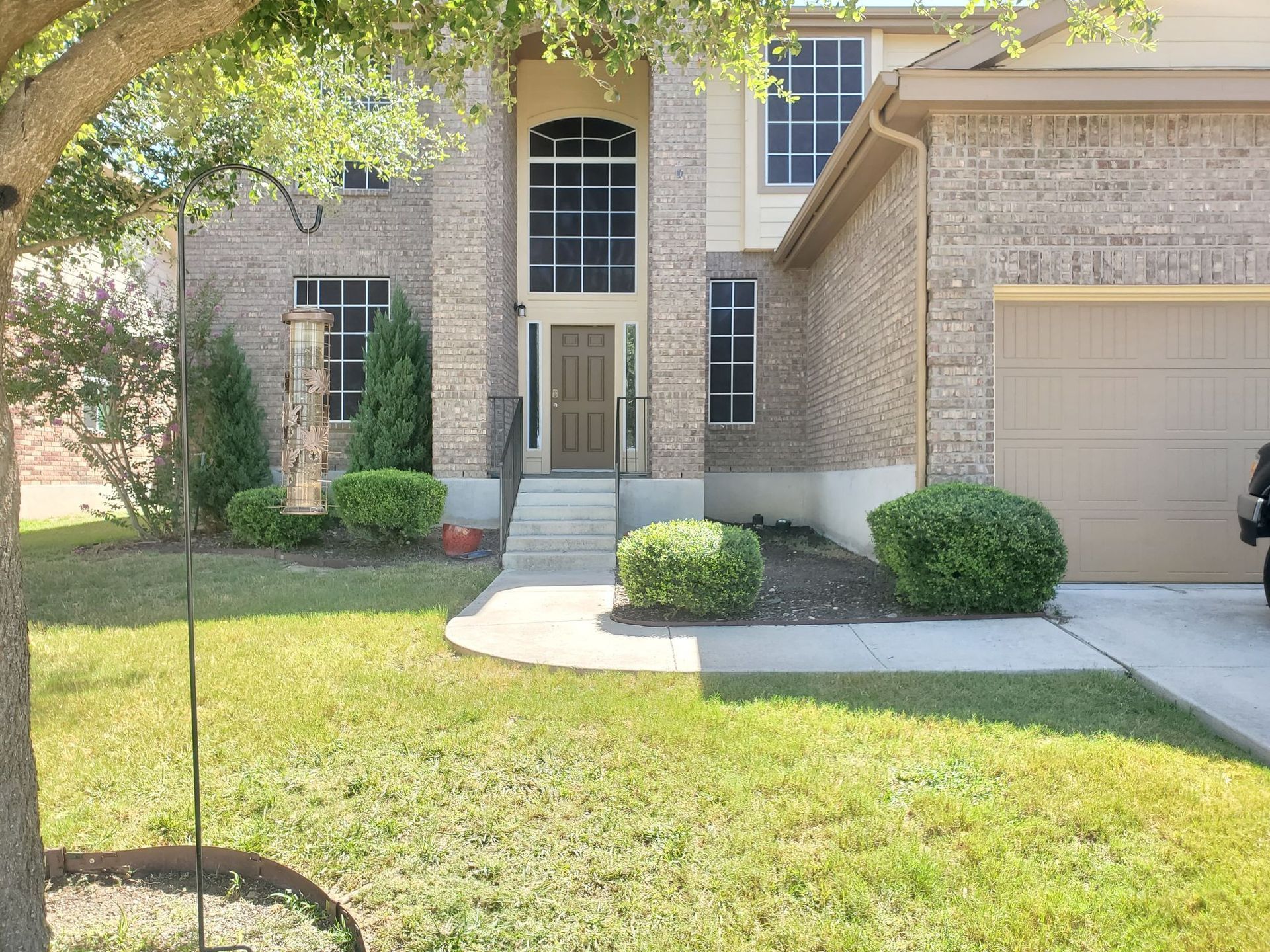 Beige brick house with front yard, curved walkway, and two-car garage. Lush green lawn and bushes.