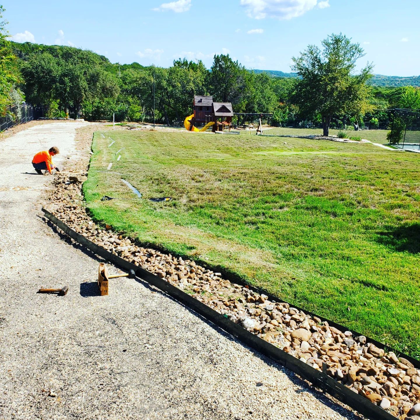 Person landscaping gravel bed next to pathway, playground in background.