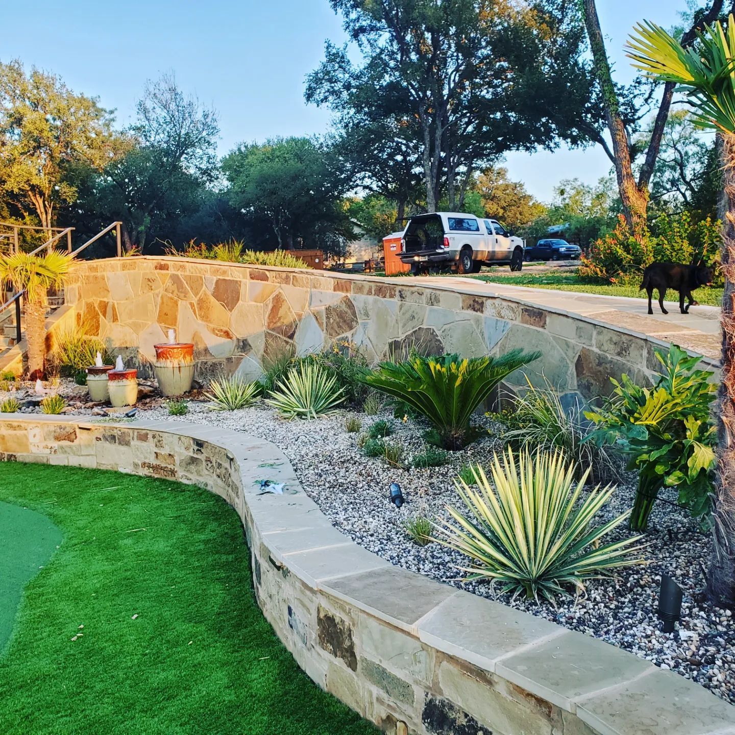 Stone retaining wall with plants, green lawn, truck parked on gravel path. Dog on the right.