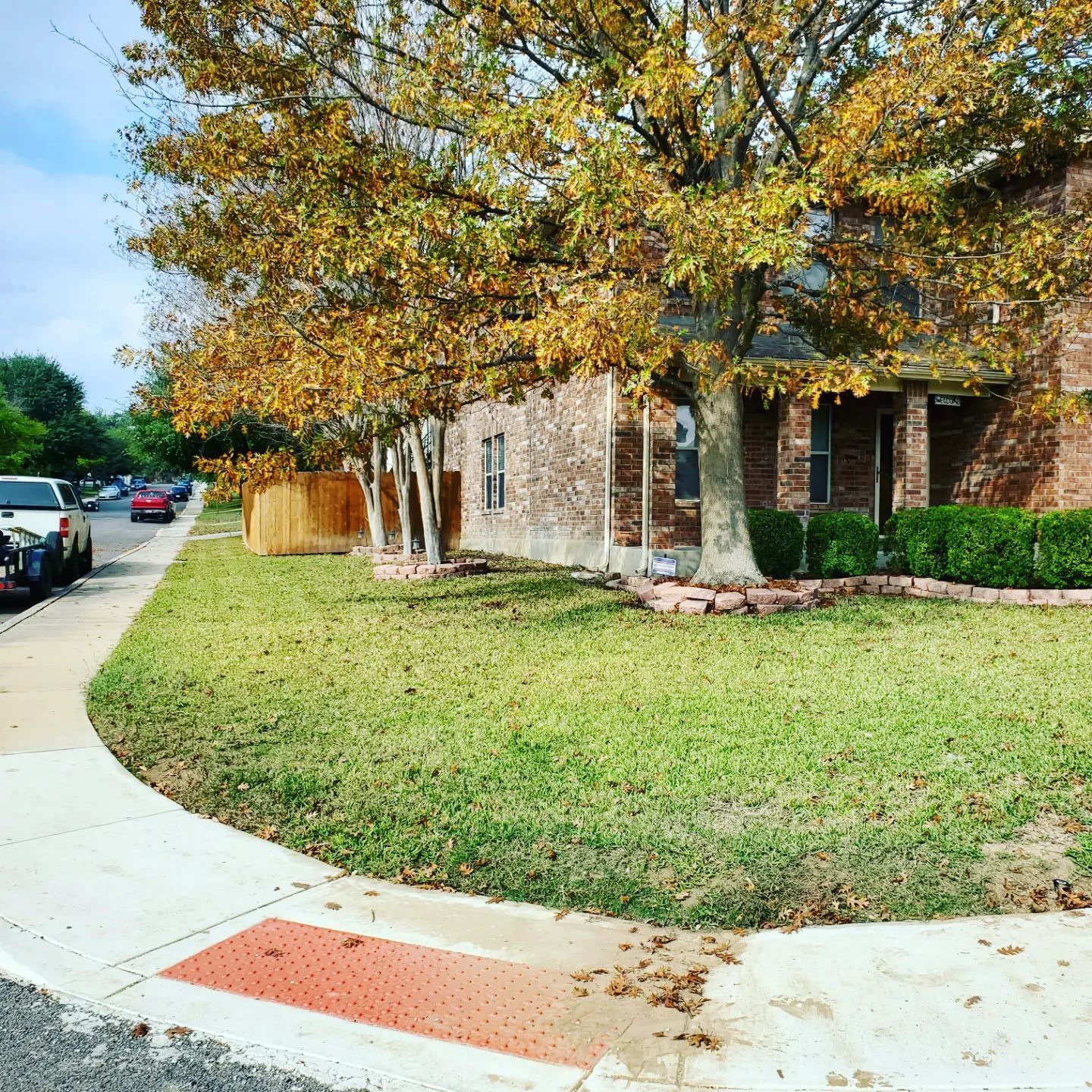 Sidewalk curves in front of a brick house with a lawn and fall foliage.