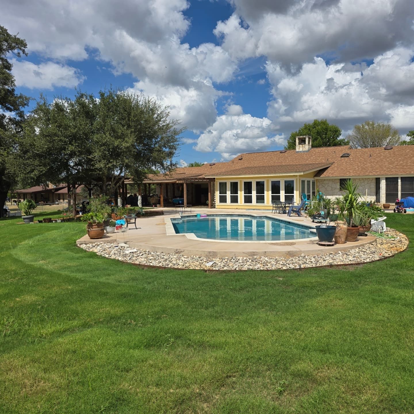 Backyard with a swimming pool surrounded by rocks, lush green grass, and a house under a cloudy sky.