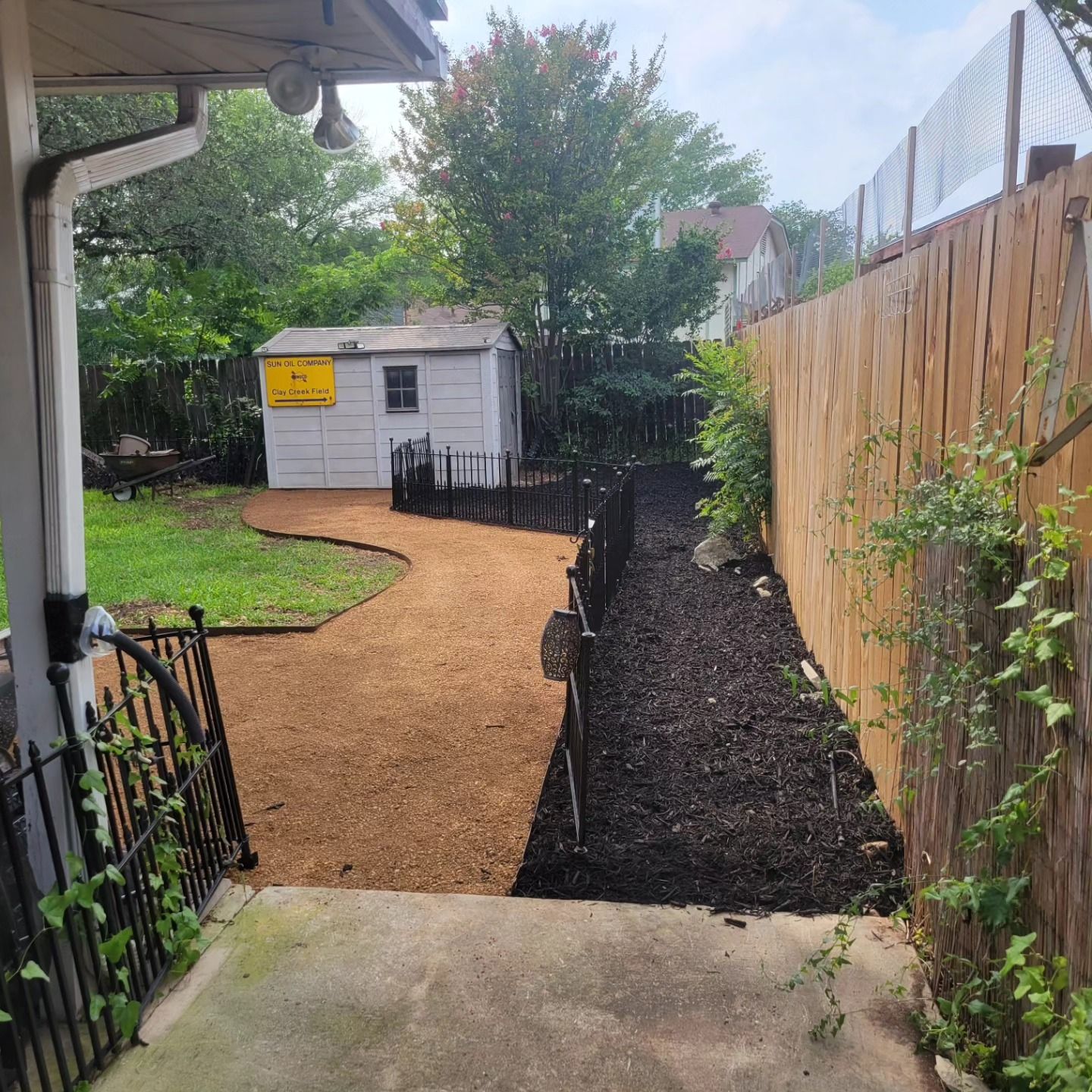 Backyard view with gravel path, shed, and wooden fence. Lush greenery and dark mulch borders the path.