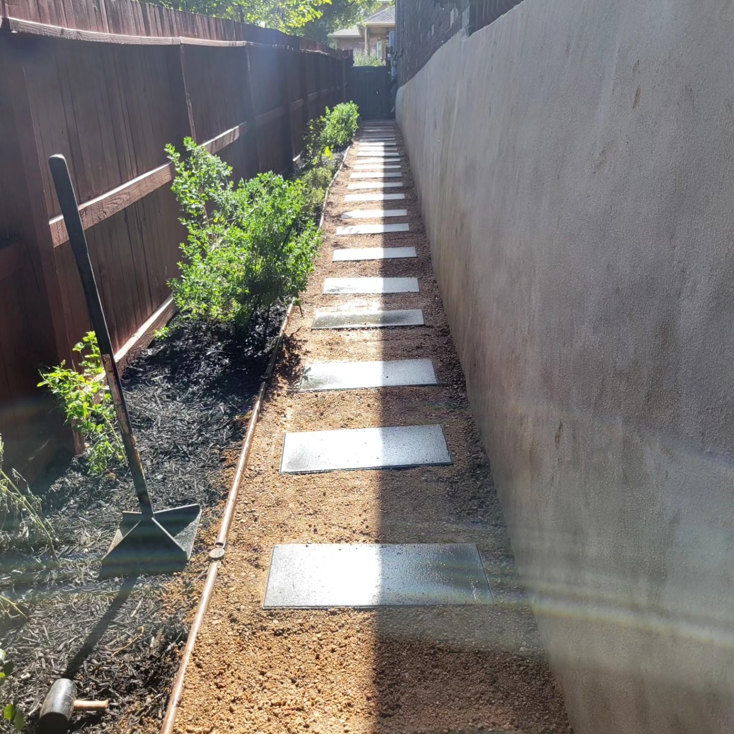 Stone path between brown fence and stucco wall, with landscaping and shovel leaning against the fence.